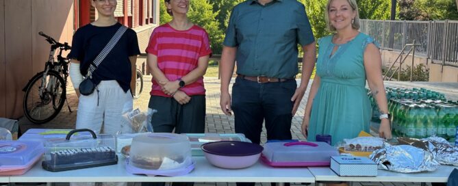 Zu sehen sind 4 Personen die hinter einer Kuchentheke stehen und freundlich in die Kamera lächeln